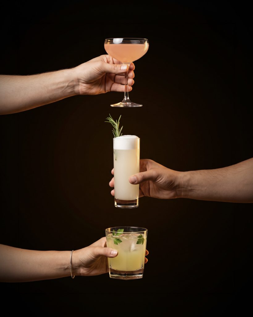 Three hands holding the three winning cocktails from the Batch 22 Cocktail Contest: Pianosa in a coupe, Buzz Fizz in a tall glass and Herb Garden in a rocks glass